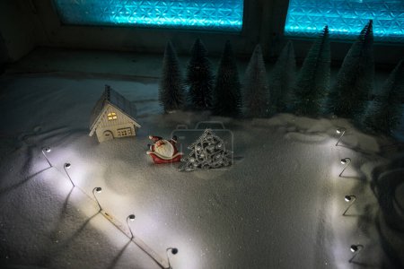 Téléchargez les photos : Noël et Nouvel An maison miniature dans la neige la nuit avec sapin. Petite maison de jouets sur neige avec des arbres et des lumières de ville bokeh sur fond. Décorations de Noël. Concentration sélective - en image libre de droit