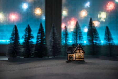 Téléchargez les photos : Noël et Nouvel An maison miniature dans la neige la nuit avec sapin. Petite maison de jouets sur neige avec des arbres et des lumières de ville bokeh sur fond. Décorations de Noël. Concentration sélective - en image libre de droit