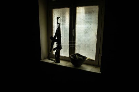Téléchargez les photos : Photo conceptuelle de la guerre entre la Russie et l'Ukraine. Arme russe et casque sur le rebord de la fenêtre la nuit. Vieille chambre flippante avec fenêtre. Explosion extérieure. - en image libre de droit