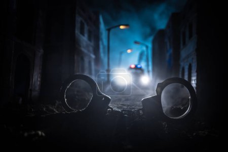 Polizeirazzia in der Nacht und Sie stehen unter Arrest. Silhouette von Handschellen mit Polizeiauto auf der Rückseite. Bild mit den blinkenden roten und blauen Polizeilichtern vor nebligem Hintergrund.