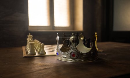 Foto de Hermosa miniatura corona en tablero de ajedrez. juego de mesa de ajedrez concepto de ideas de negocio. Figuras de ajedrez sobre un fondo oscuro con humo y niebla. Enfoque selectivo - Imagen libre de derechos