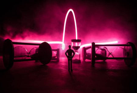 Téléchargez les photos : Concept du temps. Silhouette d'un homme debout entre des sabliers avec de la fumée et des lumières sur un fond sombre. Image décorée surréaliste - en image libre de droit