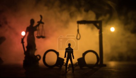 Rechtliches Recht oder Verbrechen und Hinrichtungskonzept. Miniaturen der Todesstrafe auf dem Tisch. Der Mann allein, der nachts auf die Exekution schaut. Kunstwerk-Dekoration mit Handschellen, Statue der Gerechtigkeit und Schlägel der Gerechtigkeit