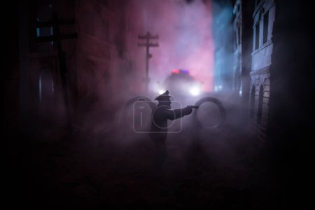 Photo for Police raid at night and you are under arrest concept. Silhouette of handcuffs with police car on backside. Image with the flashing red and blue police lights at foggy background. - Royalty Free Image