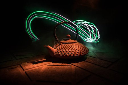Téléchargez les photos : Bouilloire arabe sur une surface en pierre sur fond sombre avec lumières et fumée. Concept de thé oriental. Concentration sélective. - en image libre de droit