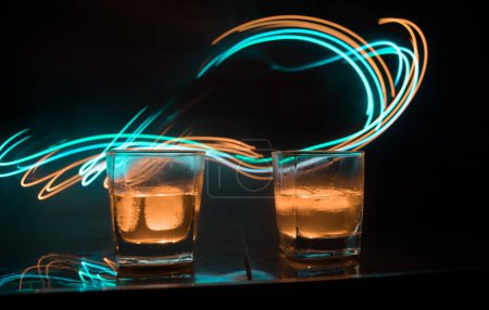 Téléchargez les photos : Concept de boisson au whisky. Verre de whisky et de glace avec lumière de couleur et brouillard sur fond de barre sombre. Concentration sélective - en image libre de droit