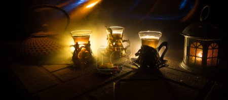Téléchargez les photos : Thé arabe en verre avec des collations orientales sur une surface en pierre sur fond sombre avec des lumières et de la fumée. Concept de thé oriental. Concentration sélective. - en image libre de droit