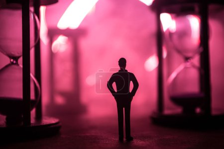 Téléchargez les photos : Concept du temps. Silhouette d'un homme debout entre des sabliers avec de la fumée et des lumières sur un fond sombre. Image décorée surréaliste - en image libre de droit