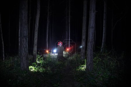Photo for A scary evil little clown wearing a dirty costume in the woods at night. Night forest, horror Halloween concept - Royalty Free Image