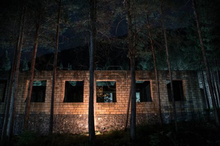 Téléchargez les photos : Ancien bâtiment abandonné dans la forêt, ruines de façade de l'usine industrielle. Printemps longue forêt de pins la nuit - en image libre de droit