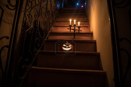 Téléchargez les photos : Horreur concept Halloween. Effrayant château abandonné. Un vieux chandelier et une citrouille d'Halloween qui brillent sur des escaliers en bois avec une porte en treillis la nuit. Décoration avec rétro-éclairage et brouillard. Concentration sélective - en image libre de droit