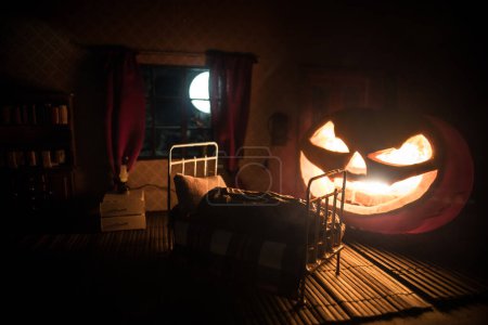 Foto de Concepto de Halloween de horror. Una casa de muñecas realista dormitorio con muebles y ventana por la noche. Hombre asustado en la cama con calabaza gigante brillante. Enfoque selectivo - Imagen libre de derechos