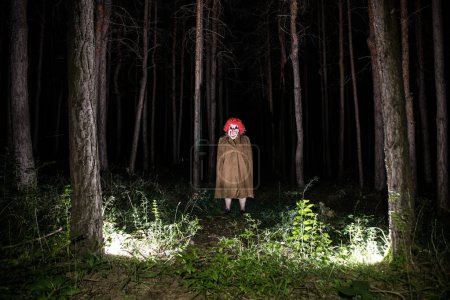 Téléchargez les photos : Un clown maléfique effrayant portant un costume sale dans les bois la nuit. Forêt nocturne, horreur concept Halloween - en image libre de droit