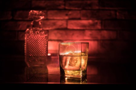 Photo for Whiskey drink concept. Glass of whiskey and ice with color light and fog on dark bar background. Selective focus - Royalty Free Image