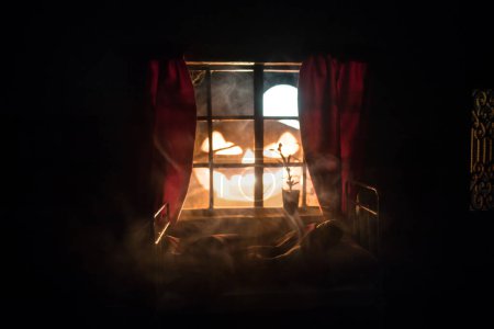 Photo for Horror Halloween concept. A realistic dollhouse bedroom with furniture and window at night. Scared man in bed with giant glowing pumpkin. Selective focus - Royalty Free Image