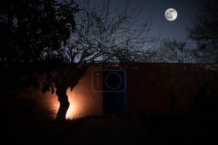 Téléchargez les photos : Pleine lune au-dessus du village tranquille la nuit. Mur orange avec porte métallique bleue la nuit. Atmosphère d'horreur hantée. Plan d'exposition longue - en image libre de droit