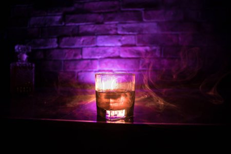 Téléchargez les photos : Concept de boisson au whisky. Verre de whisky et de glace avec lumière de couleur et brouillard sur fond de barre sombre. Concentration sélective - en image libre de droit