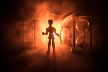 Foto de Concepto de tiempo. Silueta de un hombre parado entre clepsidras con humo y luces sobre un fondo oscuro. Imagen surrealista decorada - Imagen libre de derechos