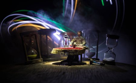 Foto de Concepto de tiempo. Hombre sentado en la mesa con reloj de arena. Reloj de arena abstracto con humo y luces sobre un fondo oscuro. Imagen surrealista decorada - Imagen libre de derechos