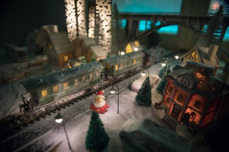 Téléchargez les photos : Miniature de scène hivernale avec maisons de Noël, gare, arbres, couverte de neige. Scène de nuit. Nouvel an dans la ville des jouets vintage. Concentration sélective - en image libre de droit