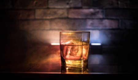 Foto de Concepto de bebida de whisky. Vaso de whisky y hielo con luz de color y niebla sobre fondo de barra oscura. Enfoque selectivo - Imagen libre de derechos