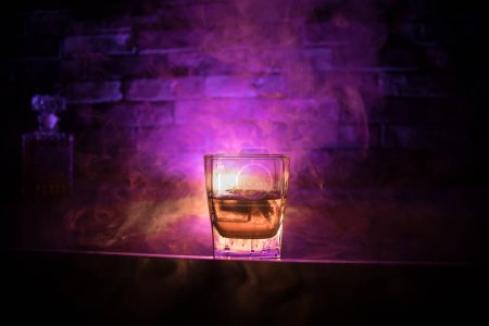 Whiskey drink concept. Glass of whiskey and ice with color light and fog on dark bar background. Selective focus