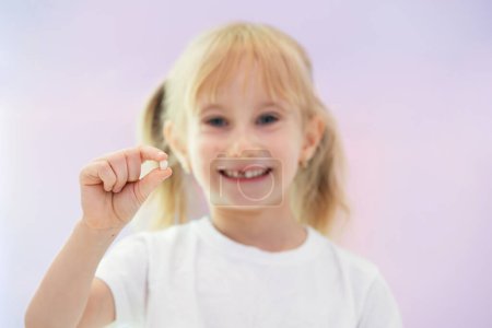 Photo for Six year old blondy smiling happy girl holding a fallen baby tooth against pink background. - Royalty Free Image