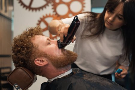 Photo for Side view of red-haired bearded man sitting in barbershop and female barber making hairdo to guest - Royalty Free Image