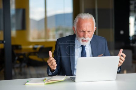 Téléchargez les photos : Bel homme âgé souriant assis à son lieu de travail confortable avec ordinateur portable à la maison, retraité mâle bavarder avec des amis dans les médias sociaux - en image libre de droit
