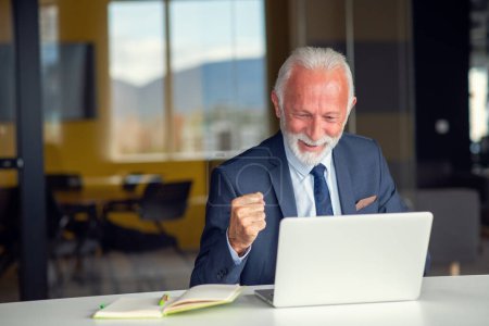 Téléchargez les photos : Bel homme âgé souriant assis à son lieu de travail confortable avec ordinateur portable à la maison, retraité mâle bavarder avec des amis dans les médias sociaux - en image libre de droit