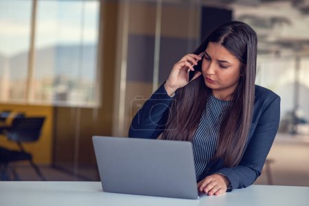 Téléchargez les photos : Femme d'affaires sérieuse parlant sur un téléphone portable au bureau, travaillant à l'ordinateur. - en image libre de droit