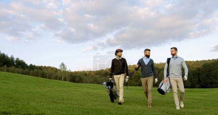 Téléchargez les photos : Three handsome men strolling with their clubs and golf bags on the golf field to start a game and talking. Outdoor - en image libre de droit