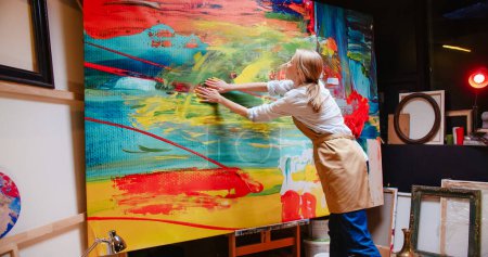 Foto de Rear of emotional Caucasian young innovative professional woman artist working in workshop at night and drawing on big canvas with her hands creating abstract art piece, creative work, hobby concept - Imagen libre de derechos