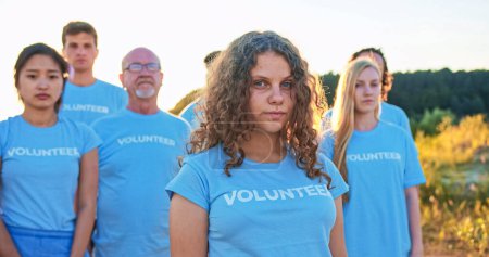 Téléchargez les photos : Jolie jeune fille avec un groupe de militants écologistes en arrière-plan. Belle bénévole caucasienne regardant la caméra et disant : Je suis bénévole. Volontariat, conservation de l'environnement. - en image libre de droit