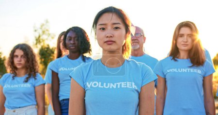 Téléchargez les photos : Jeune belle femme avec un groupe d'éco-activistes en arrière-plan. Jolie bénévole asiatique regardant la caméra et disant : Je suis bénévole. Volontariat, conservation de l'environnement. - en image libre de droit