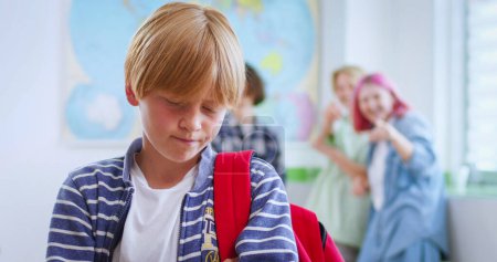 Trauriger gemobbter Junge, der im Klassenzimmer steht, während drei Mitschüler hinter ihm spötteln. Konzept der Diskriminierung und negativen Kommunikation in der Gesellschaft.