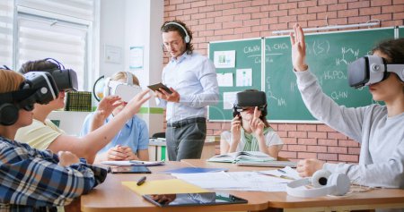 Photo for Group of young pupils in VR headsets sitting together at desk while male teacher with digital tablet in hands explaining education program. Modern technologies during interactive lesson at school. - Royalty Free Image