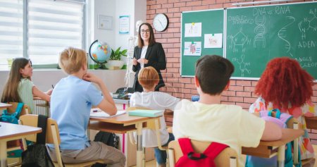 Cheerful Caucasian young female teacher of anatomy or biology talking to class and explaining human DNA to pupils. At school. Education concept. Indoors. Teaching. Lection about body.-stock-photo