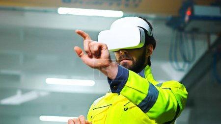 Photo for Close up of Caucasian male young engineer in headset and yellow uniform standing in factory and having virtual reality experience. Engineering concept. Indoors. Man in VR goggles moving hands in air. - Royalty Free Image