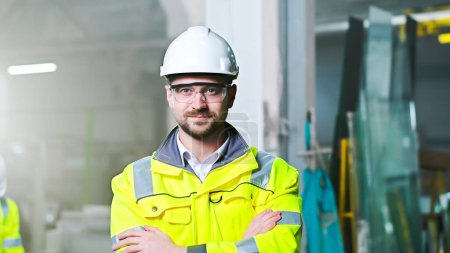 Portrait of handsome Caucasian man in helmet and uniform standing at plant manufactoring and taking on goggles. Indoors. Young worker at factory in hardhat and protectional glasses. Protection.