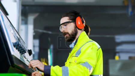 Close up of Caucasian workman in headphones and yellow uniform pushing buttons on controlling point and running michine at plant. Inside. Technician man in goggles at controller computer in factory.