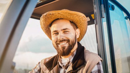 Close up of Caucasian handsome happy young man farmer in hat sitting in cabin of tractor and smiling to camera. Countryside work concept. Portrait of attractive cheerful male driver having rest.