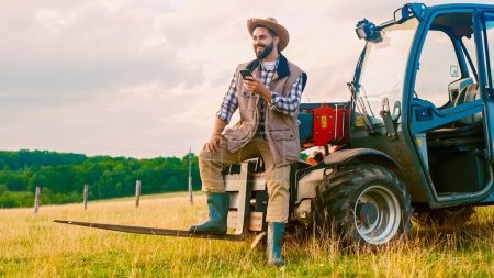 Caucásico joven granjero guapo en sombrero de pie en el tractor, utilizando el teléfono inteligente y descansando en el campo. Concepto de trabajador rural. Feliz macho descansando y enviando mensajes de texto por teléfono mientras chatea. Exterior.