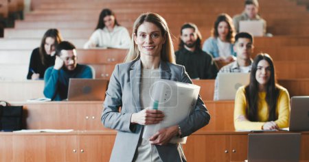 Photo for Portrait of beautiful Caucasian female professor at high school standing at classroom with papers and smiling to camera. Pretty cheerful woman teacher at college or University. Students behind. - Royalty Free Image