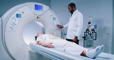 Multicultural doctor is pushing buttons at CT scanner. Patient is finished magnetic resonance imaging. African american doctor performed MRI scanning procedure. Ethnic diversity.