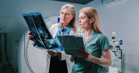 Photo for Female doctor and nurse standing at equipped tomography room. Doctor is holding scans and consulting with colleague. Nurse hold folder and listen to doctor carefully. Analizing of examination results - Royalty Free Image