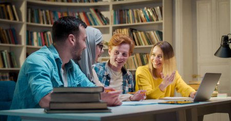 Photo for Mixed-races young males and females friends sitting at table in library, talking cheerfully and learning. Men and women brainstorming over studying project in bibliotheca. - Royalty Free Image