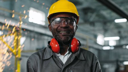 Photo for African american engineer in protective clothes. Worker stared and posing at construction site. Employee in googles and headphones smiling at camera on background of welding sparks plying. - Royalty Free Image