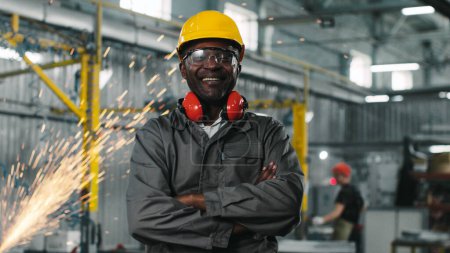 Photo for African american employee stared, smiling and posing for camera with crossed arms. Male engineer in googles and headphones at manufacture background where welding sparks flying. - Royalty Free Image