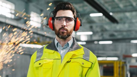 Photo for Industrial engineer in protective headphones wearing safety vest works in heavy manufacturing factory. Bearded builder is looking straight at camera. Worker at background of welding sparks flying. - Royalty Free Image